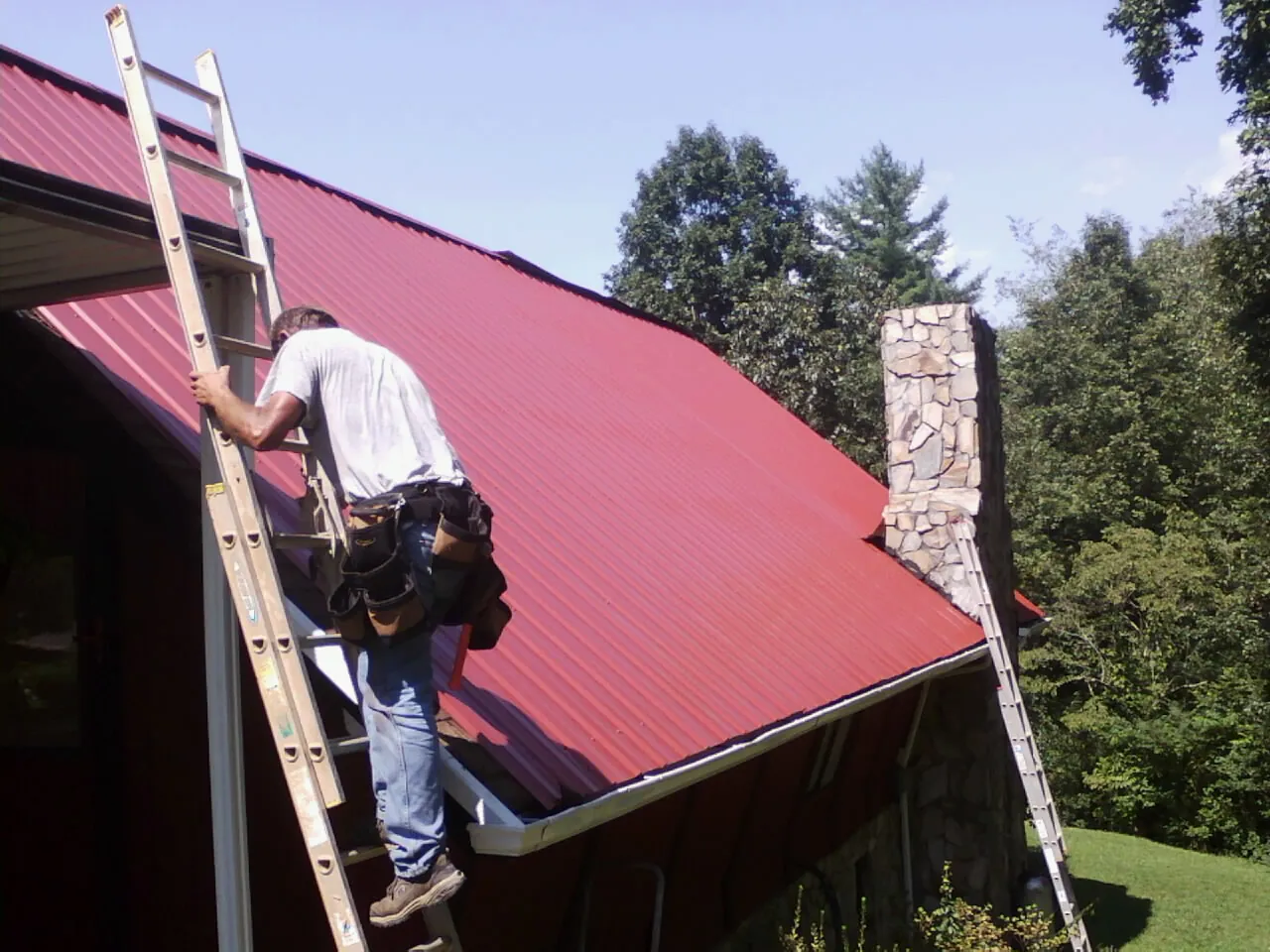 Roofer on a ladder beside a red metal roof and stone chimney — Soffit & Fascia Repair by Roofing Brothers in Dillsburg